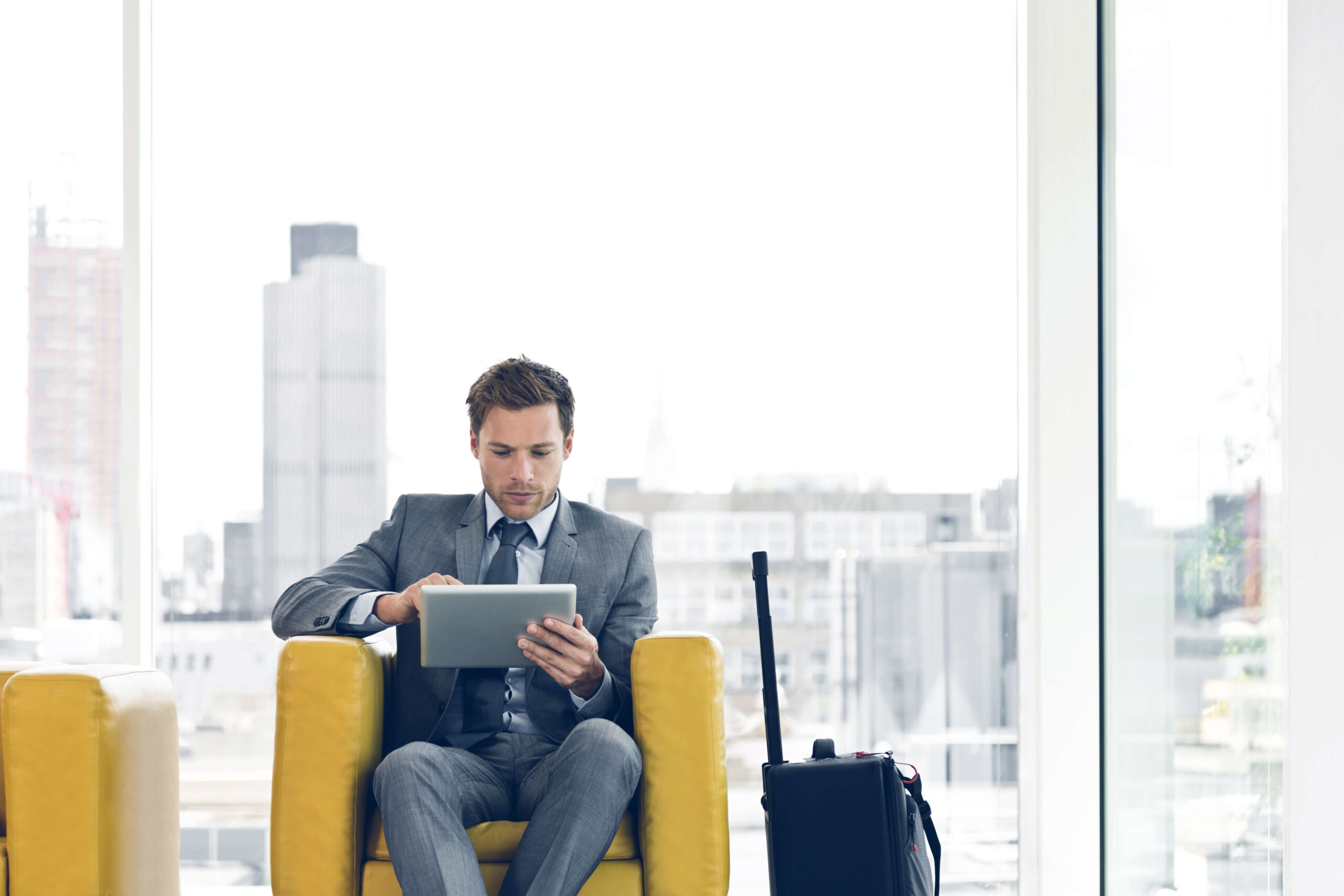 Man with suitcase on iPad sitting in yellow chair.