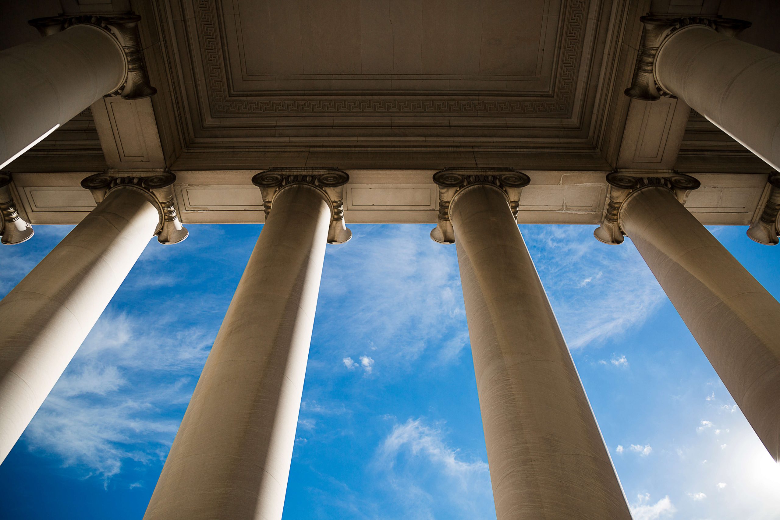 Smooth ionic columns holding a ceiling seen from a low perspective backed by a blue sky with fluffy clouds